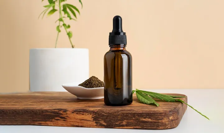 A dark glass dropper bottle on a wooden board next to a small dish of seeds and a cannabis leaf, with a potted plant in the background.